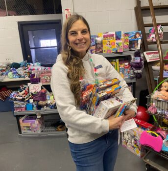 Volunteer and local teacher jane Harper, a white woman with long brown hair, a gray sweatshirt, and light blue jeans, holds an armload of puzzles as she helps someone shop for a child in need.