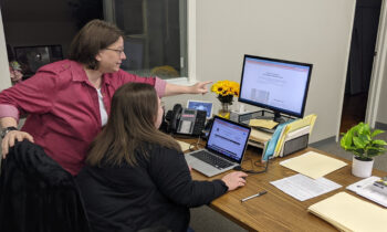 Jenni points at something on a computer monitor while talking to Katie. Jenni is a white woman with glasses, a serious expression, chin-length brown hair, a white shirt with a red button-down shirt over top with the sleeves rolled up.