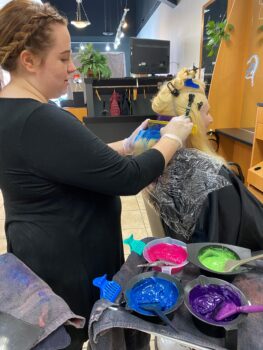 Elyssa, a young white woman with light brown hair in a braid wrapped around her head, freckles, and a black shirt, puts hair color in a client's blonde hair. colors of bright green, pink, blue and purple are next to her.