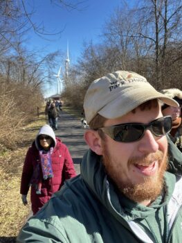 James Anderson, a tall white man with a white ballcap, sunglasses, a red beard and a green hoodie, leads walkers bundled up in coats, hats, and scarves on a winter hike. 