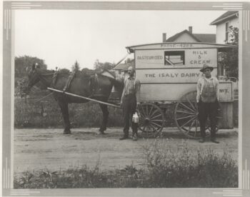 The black-and-white photo shows two men standing next to a wagon with "the Isaly Dairy Co. - Pasteurized milk & Cream; Phone - 4289" on the side. It is bulled by a black horse. The two white men wear long-sleeved, collared, button up shirts and dark pants. One man holds a wire basket with two milk bottles in it. One man has a hat and the other has a mustache. the dirt road is surrounded by wildflowers. Several houses are visible in the background.