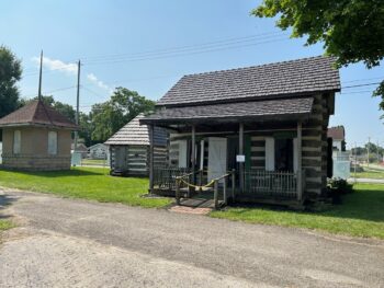 The log cabin is in the foreground with a small porch. Teh log smokehouse, a smaller building, is next to it. A brick ticket booth for the urban interurban electric railway is o nthe far left.