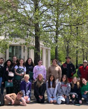 16 high school students in the TRIO program pose on the campus of Ohio University. There are green trees and a building in the background. 