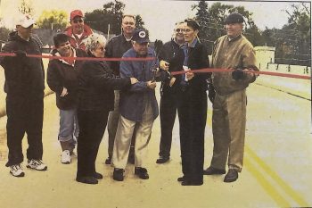 A small group of people look on as a red ribbon is cut. 