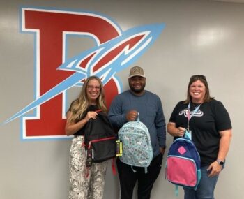 A black man and two white women hold backpacks in front of the Ridgedale Rockets logo.
