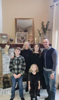 The family smiles in a group picture in front of a mantle. They have one boy and two girls. 