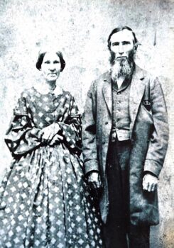 A black-and-white photo fof an unsmiling couple. Mary Ellen has brown hair pulled into a bun, a patterned dress with a lace color, long sleeves, and a full skirt. Hartman has short dark har, sharp cheeckbones, and a long beard with white in teh front. He ears a long overcoat, and has a vest. 