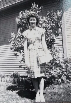 Mary Ellen is shown as a young woman with a white hat, curly short brown hair, a smile, a plaid drewss with ruffled short sleeves and peplumb at the waist under a belt. The skirt goes just below her knees. She has white dress shoes with black toes. She carries a white purse and wears short white gloves. 