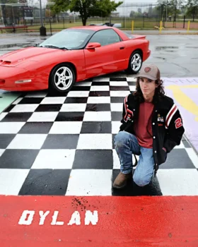 Dylan wears a Pleasant letter hacket in front of a parking space that says 'Dylan' while a red sports car is in the background.