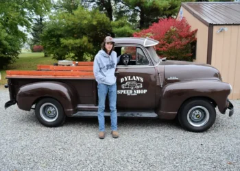Dylan stands next to a small classic brown truck and pets a black dog sitting inside. The door has the logo: Dylan's Speed Shop.