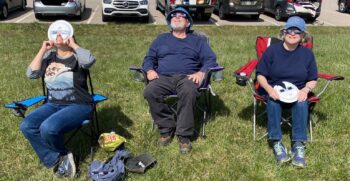 Three people sit in camping chairs and look at the partial solar eclipse. 