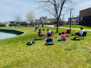 Dozens of people sit in chairs and on the green grass. Flowering trees and the campus pond are visible. Some people are standing and looking at the partial solar eclipse with safety glasses. 