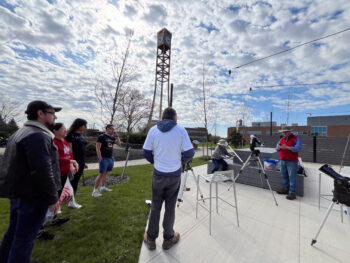 People wait to view the solar eclipse through telescopes on campus. The campus bell tower is visible in the background.