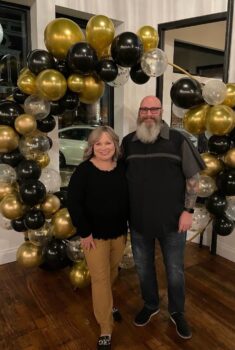 A couple smiles in front of a black, gold, and white balloon sculpture.