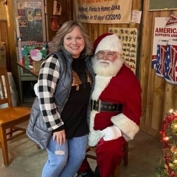 Jodi, a white woman with light brown wavy hair and a smile, sits on the lap of Santa at a fundraiser for the Christmas Clearinghouse.