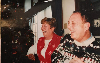 A historical photo of Frey's parents smiling and laughing in front of a Christmas tree while wearing Christmas sweaters.