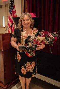 Jodi smiles and holds her gold award and a bouquet of flowers. 