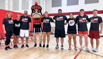 The Pleasant corporate dodgeball team and the Pleasant Spartan Mascot smile in a gyn. 