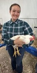 Meleah smiles while holding a large white chicken. She has her brown hair back in braids. She wears a green, blue and white plaid long-sleeved shirt, dark jeans and brown boots. 