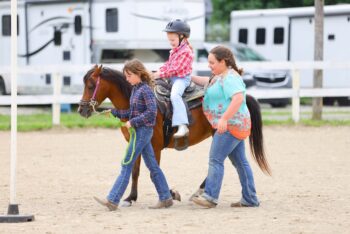 A small girl wears a ribbon and rides a pony while two other girls walk along side. 