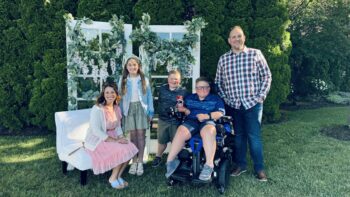 The family smiles outdoors. One boy is in a motorized wheelchair. Both boys have Duchenne's muscular dystrophy.