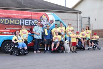 Chris Andrews, the Fox family, and others including three boys in wheelchairs with Duchenne muscular dystrophy, smile next to a van covered with the 'My DMD Hero' logo and info. 