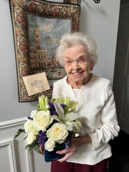 A white-haired woman with glasses and a smile holds a bouquet of flowers. 