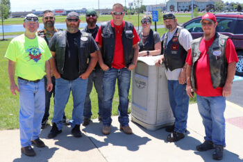 Seven men and one woman in sunglasses, black leather jackets and jeans smmile together. 