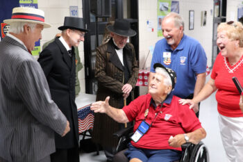 A veteran in a wheelchair smiles and holds out his hand to a man playing President Warren G. Harding. Two other volunteers in historical clothes smile. Behind the veteran, two supports smile. 