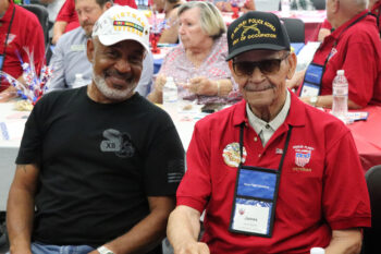 A tall black man with a black "Miitary police Korea" hat, a red "Honor Flight Columbus Veteran" shirt, glasses and a mustache gives a shy smile. His nephew, a black man with a smile and a white beard, sits next to him. The nephew wears a white Vietnam Veteran Hat and a Black military T-shirt.