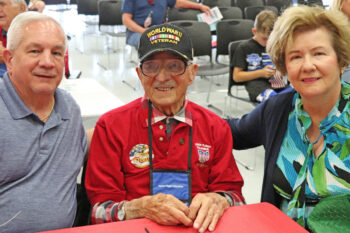 A 100-year-old white man with a black ballcap with the name "World War II Veteran"  and a red "Honor Flight Columbus" shirt smiles. His adult daughter sits on one side and his adult son sits on the other. 