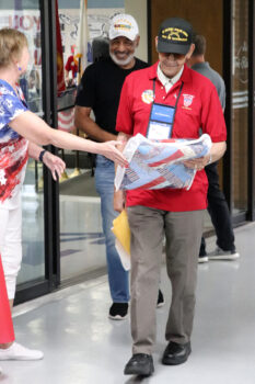 A tall black man walks independently. The veteran smiles while receiving a patriotic quilt from a woman in red, white and blue clothing. His nephew smiles in the background. 