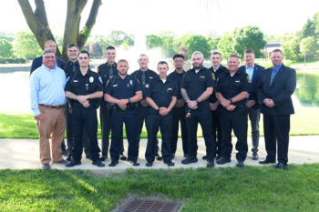 10 graduates in uniform and four instructors pose in front of the campus pond. 