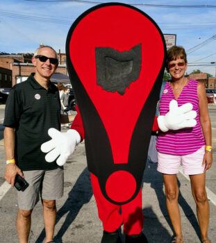 The couple stands on either side of a life-sized exclamaation mark with an outline of Ohio in the iddle and white gloved hands. 