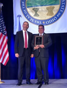 Two men smile next to a flag. Greg Perry holds a plaque. 