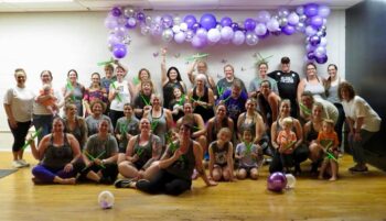 A group of about 3 dozen men, women and children smile in a dance studio. Purple balloons are in the background.