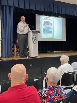 A man presents with a photo of his book behind him. Audience members sit in the foreground. 