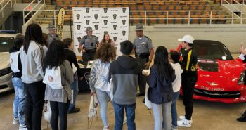 A trooper speaks to a group of students. vehicles and other highway patrol officers are at a table with info. 