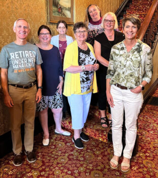 7 people smile inside the historic office of the Marion Community Foundation. Dean, a white man with short white hair and a smile, wears a gray T-shirt that states: "Retired; Under new management; see wife for details."