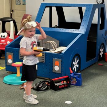 A young girl smiles while wearing goggles and holding a toy drill. She has a tool box and is working on a wooden large car at the Explore-It-Torium.