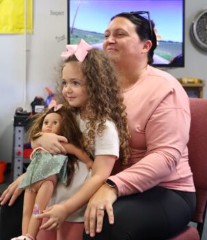 A mother holds her daughter, who has long brown curly hair with a pink bow, who is holding her doll with long brown hair and a pink bow. They are sitting at storytime at the Explore-It-Torium. 