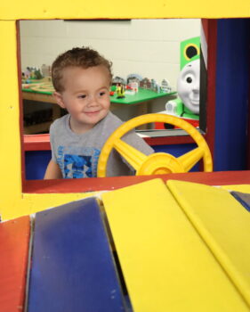 A toddler smiles while driving a wooden train at the Explore-It-Torium. A train table and giant Percy figure are visible in the background.