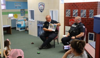 Two uniformed officers read to a circle of children at the Explore-It-Torium. Behind them are a play police station, fire department and hospital. 