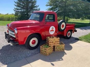 Three crates of different colored apples are stacked next to a red 1954 Ford Truck with the Country Road Farm logo on the door.