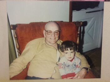 An older man with brown glasses smiles while holding a toddler with short brown hair and brown eyes. 