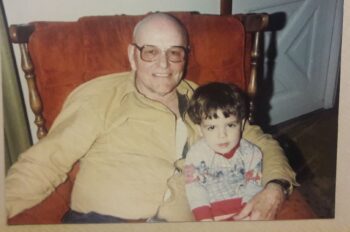 An older man with brown glasses smiles while holding a toddler with short brown hair and brown eyes.