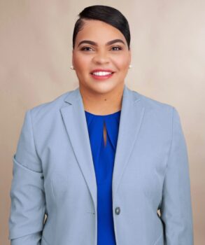 A bvlack woman smiles. She has short, sleek hair, pearl earrings, a light blue suit jacket over a bright blue blouse.