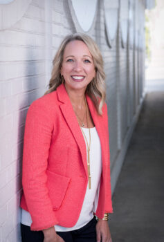 A smiling woman with long, wavy blonde hair, a smile, and a pink suit jacket over a white bouse with gold necklaces. 