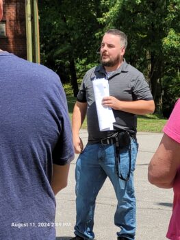 Cody is a white man with short brown hair peppered with gray, a brown beard, a gray polo and jeans. People are listening to him speak in the foreground. 