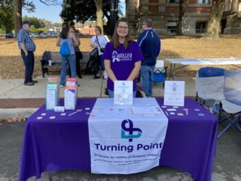 A smiling young woman in a purple shirt stands behind a table with info for turning point. 